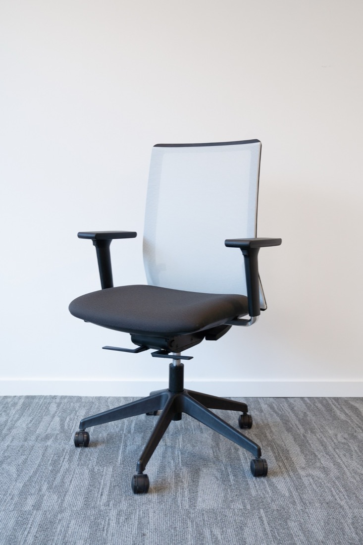 An office chair photographed against a plain white wall on grey carpet, clean, empty, shot at seat height with nothing else in frame.